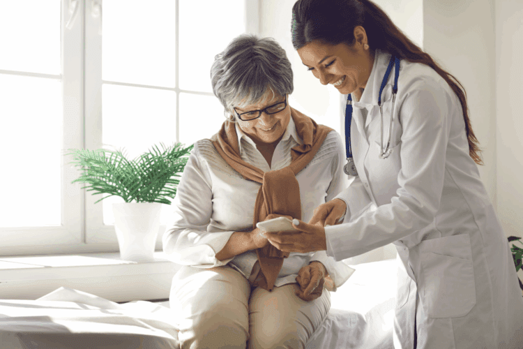 A doctor shows a smartphone to an older woman, who is seated and smiling, in a bright medical office with a plant on the windowsill.