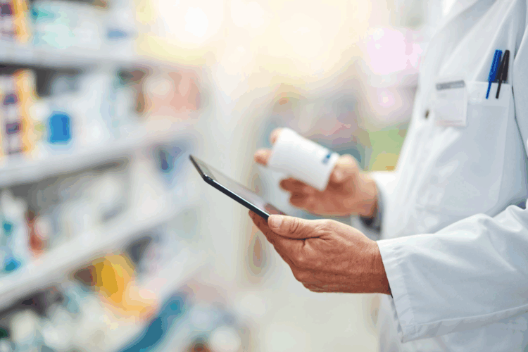 A pharmacist in a white coat holds a medication bottle in one hand and uses a tablet in the other while standing in a pharmacy aisle.