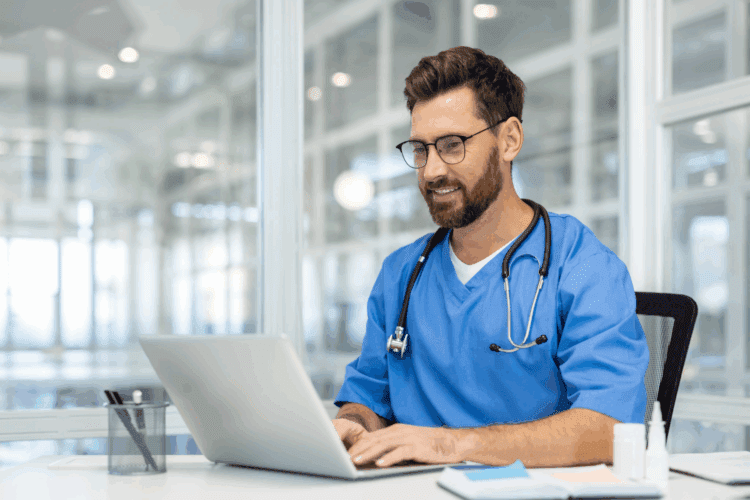 A male healthcare professional wearing blue scrubs and a stethoscope sits at a desk, typing on a laptop in a modern medical office.