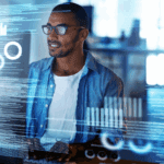 A man wearing glasses and a blue shirt works at a computer with transparent digital graphs and data visualizations overlaid in front of him.