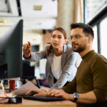 A woman standing points at a computer monitor while a man sits at a desk using the keyboard in a modern office setting.