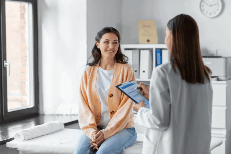 A woman sits on an exam table in a doctor's office, smiling, while a healthcare professional reviews a clipboard.