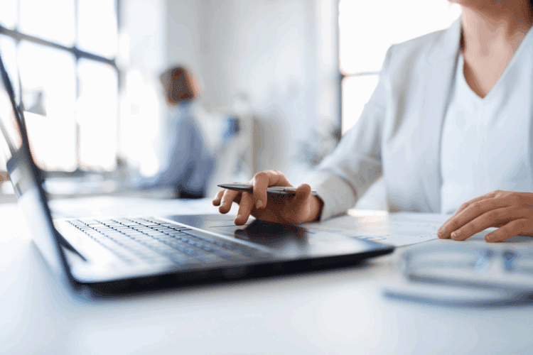 Person in business attire using a laptop and holding a pen at a desk, with another person working in the blurred background.
