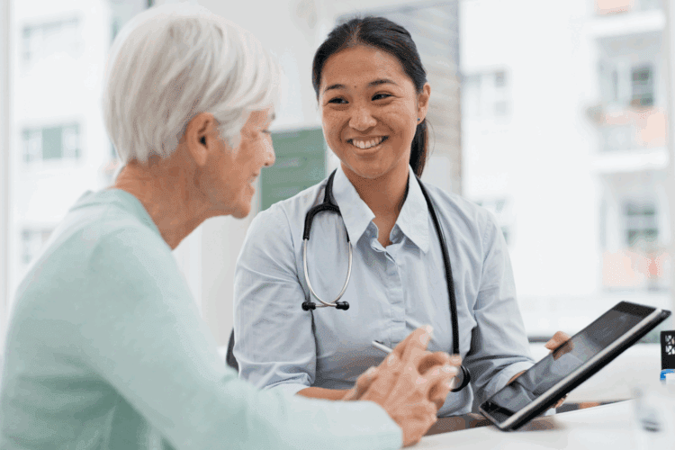 A healthcare professional with a stethoscope smiles while discussing information on a tablet with an older woman at a medical office.