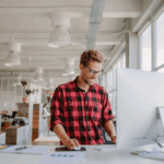 A man in a red plaid shirt stands at a desk in a modern office, working on a computer with papers and graphs nearby.