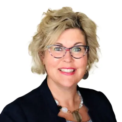 Woman with short blonde hair and glasses, smiling, wearing a dark blazer, a necklace, and earrings, against a white background.