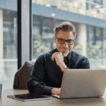 A man with glasses and a goatee sits at a desk in an office, working on a laptop. A tablet and a mug are also on the desk. Large windows show an urban background.
