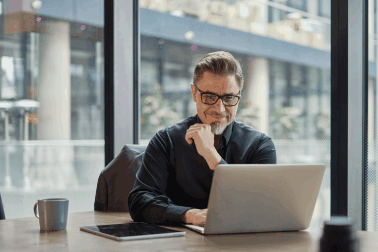 A man with glasses and a goatee sits at a desk in an office, working on a laptop. A tablet and a mug are also on the desk. Large windows show an urban background.