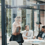 Five people in business attire discuss AI-readiness around a table in a modern office filled with natural light and large glass windows.
