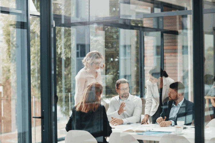 Five people in business attire discuss AI-readiness around a table in a modern office filled with natural light and large glass windows.