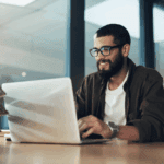 Man with glasses and a beard working on a laptop at a desk in a modern office setting.