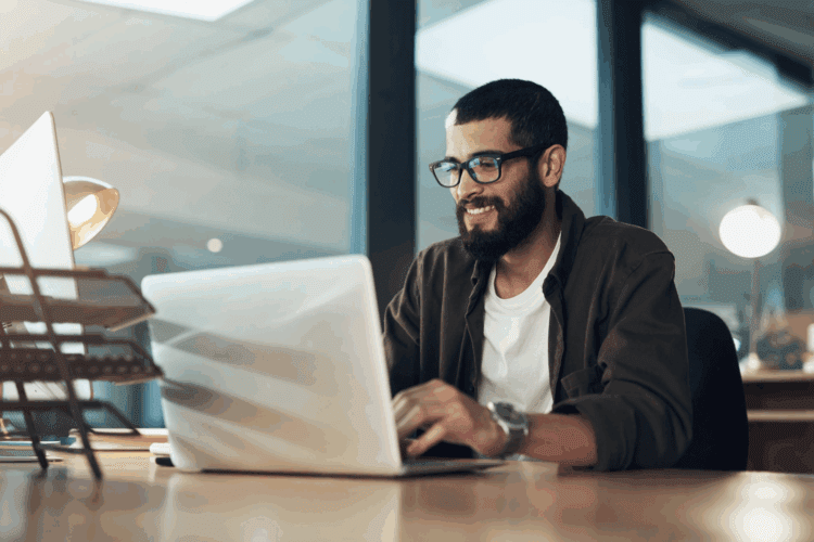 Man with glasses and a beard working on a laptop at a desk in a modern office setting.