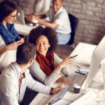 Three people sit at a desk working together on computers, conversing and smiling in a modern office space. Two more people work in the background.