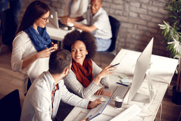 Three people sit at a desk working together on computers, conversing and smiling in a modern office space. Two more people work in the background.