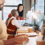 Three people work at desks in a modern office. One person stands and points at a computer screen while another sits and observes. Coffee cups and papers are on the desks.