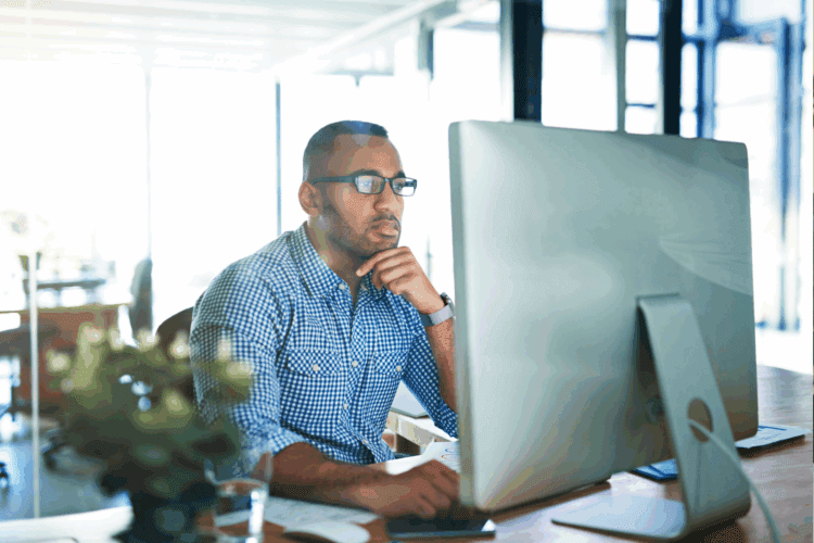 A man wearing glasses and a checkered shirt sits at a desk, looking at a large computer monitor in a bright, modern office.