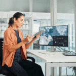 A woman sits at a desk examining a tablet, with a computer monitor displaying maps and data, in a modern laboratory or office environment.