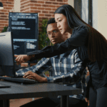Two people in an office look at a computer monitor; one is seated typing, while the other stands beside and points at the screen. Computer code is visible on a monitor in the background.