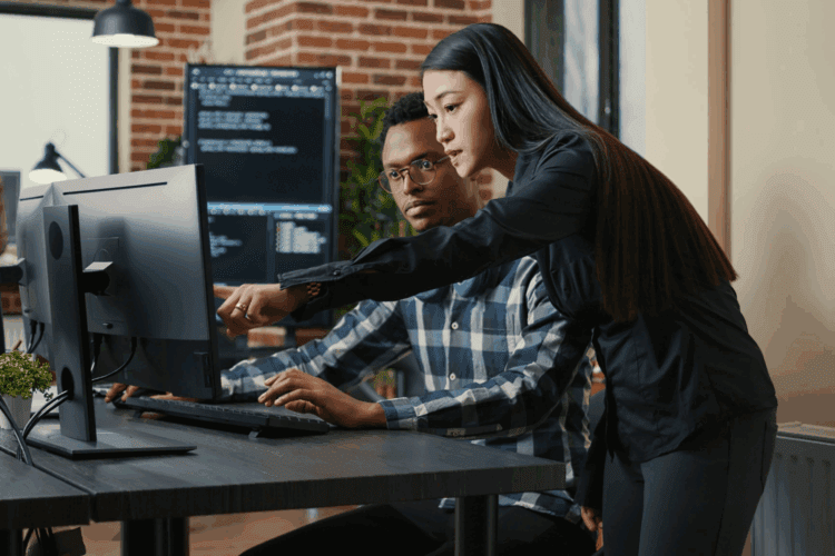 Two people in an office look at a computer monitor; one is seated typing, while the other stands beside and points at the screen. Computer code is visible on a monitor in the background.