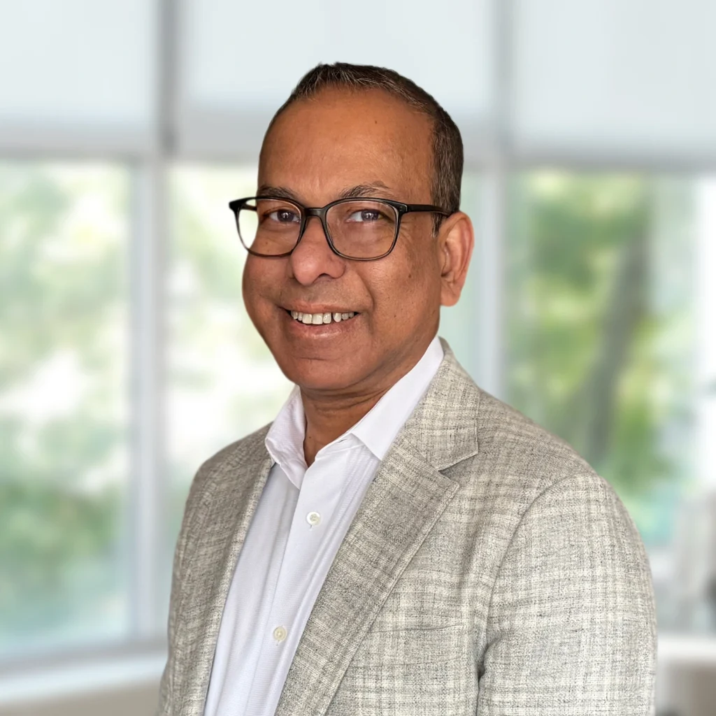 A man wearing glasses, a light grey checked blazer, and a white shirt smiles while standing indoors in front of large bright windows.