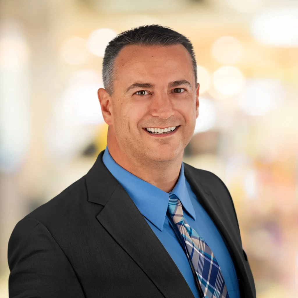 Man in a dark suit, blue shirt, and plaid tie smiling at the camera with a blurred, light-colored background.