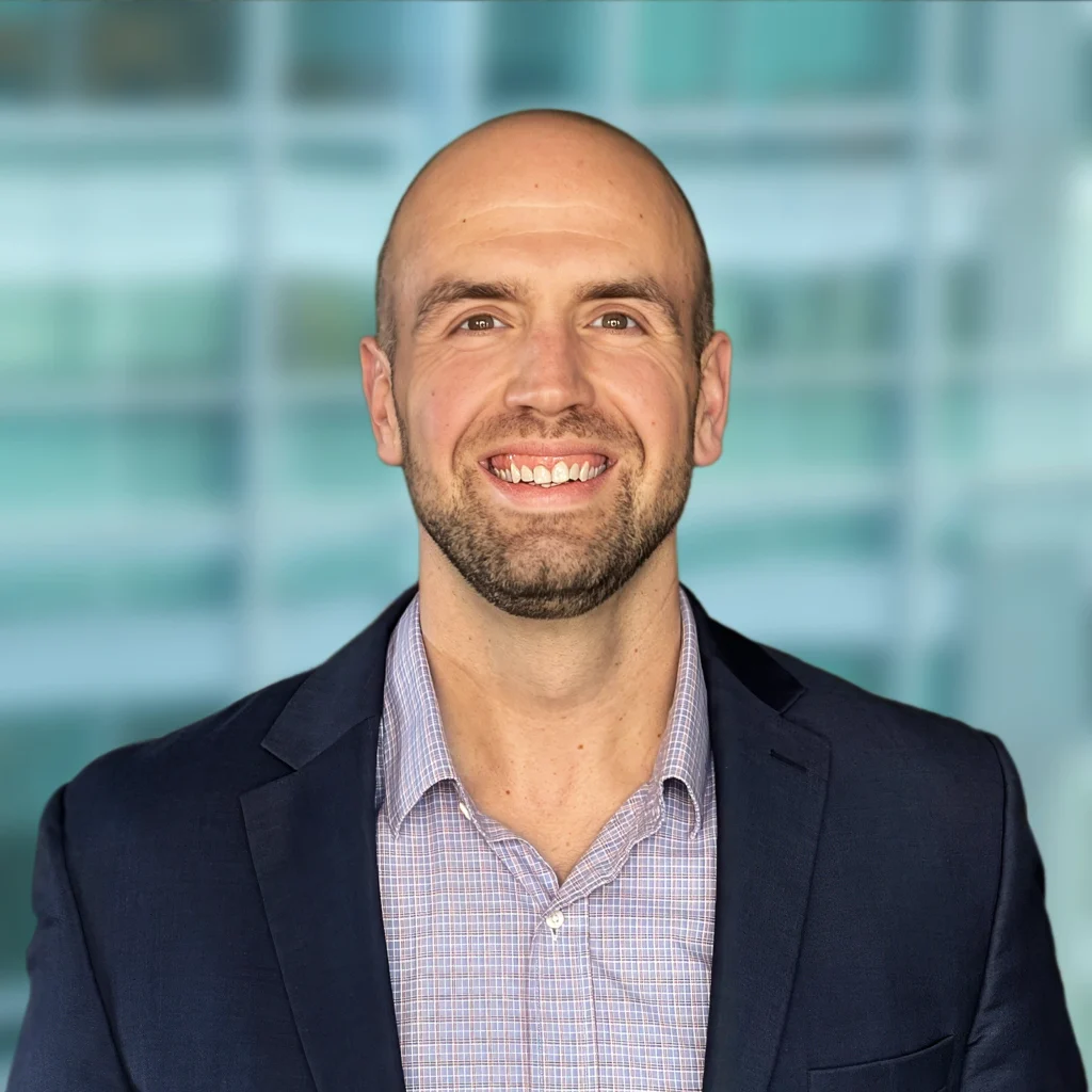 A man with a shaved head and trimmed beard, wearing a checked shirt and dark blazer, smiles in front of a blurred office background.