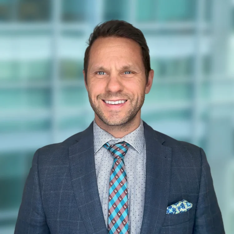 A man in a grey checked suit, patterned shirt, and blue patterned tie smiles at the camera with a blurred office background.