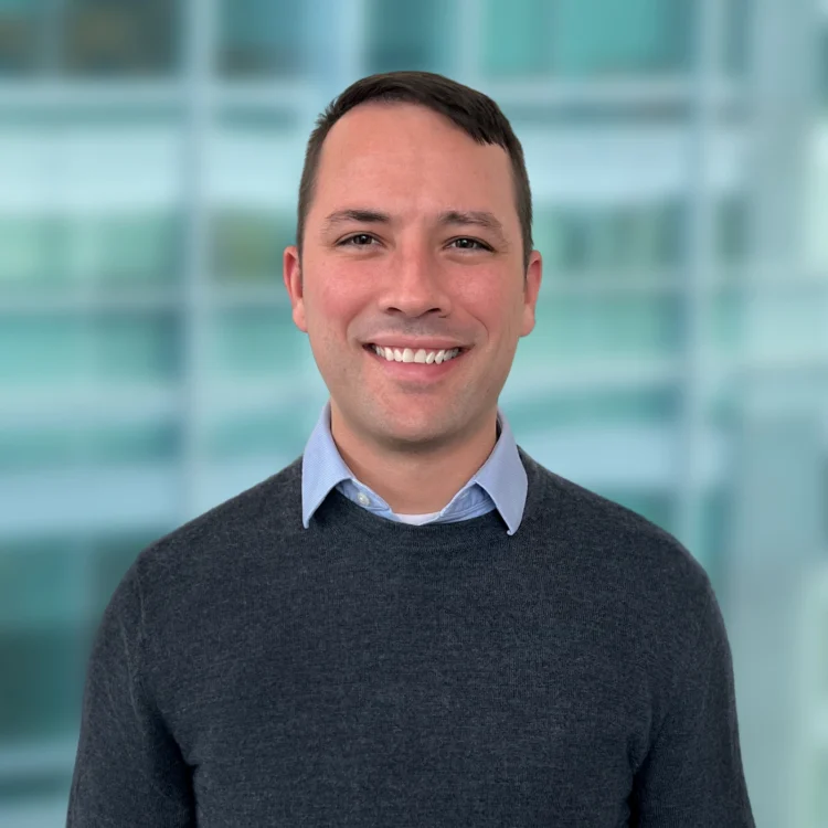 A man with short dark hair is smiling, wearing a light blue collared shirt under a dark sweater, standing in front of a blurred glass window background.