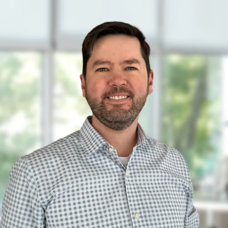 A man with short dark hair and a beard, wearing a checked shirt, stands in front of a blurred window background with greenery visible outside.