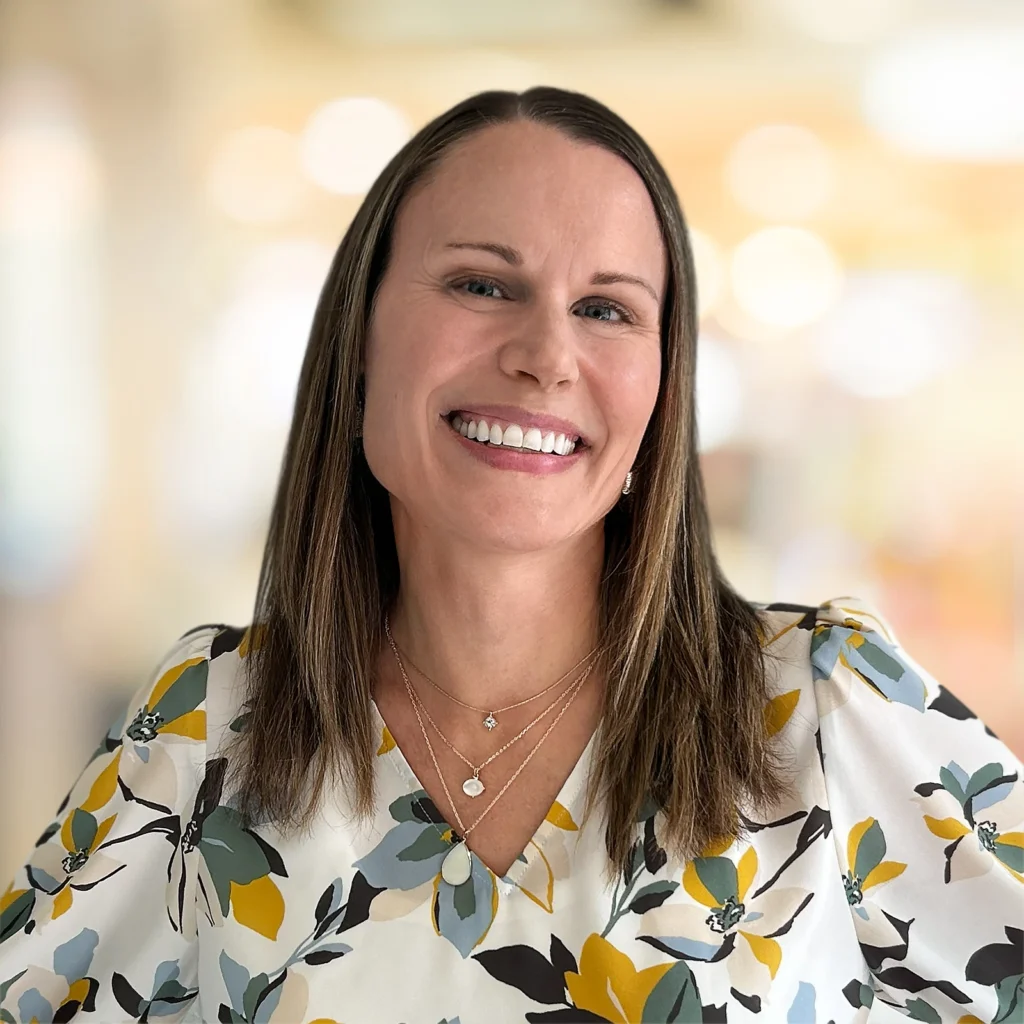 A woman with straight brown hair, wearing a floral blouse and layered necklaces, smiles at the camera against a blurred indoor background.