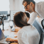 A man leans over to speak to a woman working on a laptop at a desk in an office setting, with colleagues in the background.