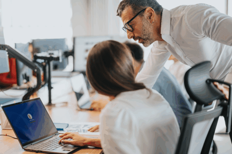 A man leans over to speak to a woman working on a laptop at a desk in an office setting, with colleagues in the background.