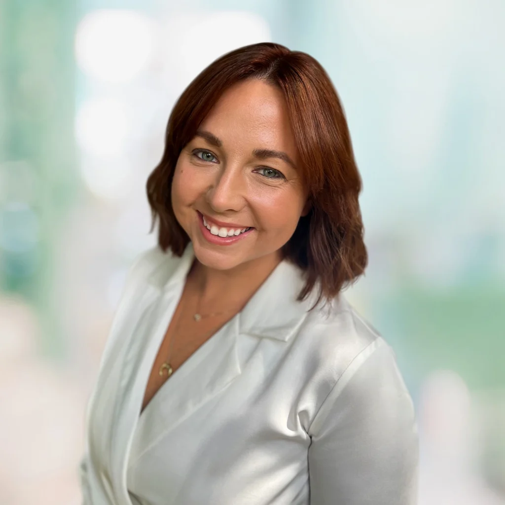 A woman with shoulder-length brown hair, wearing a white blazer, smiles at the camera against a softly blurred light green background.