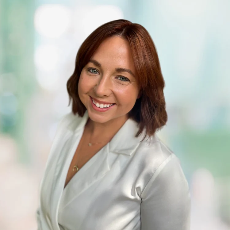 A woman with shoulder-length brown hair, wearing a white blazer, smiles at the camera against a softly blurred light green background.