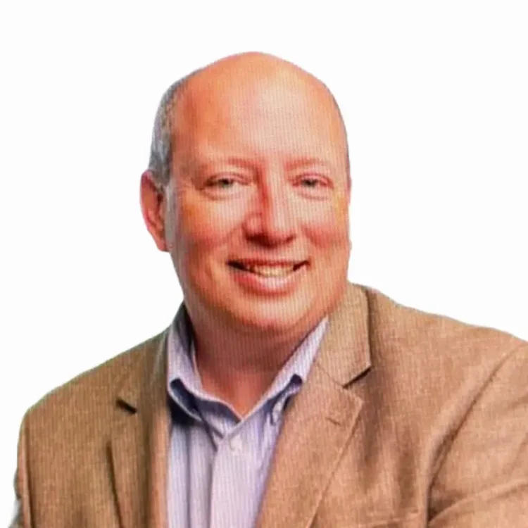 A middle-aged man with a bald head wearing a beige blazer and light blue collared shirt, smiling, in front of a plain white background.