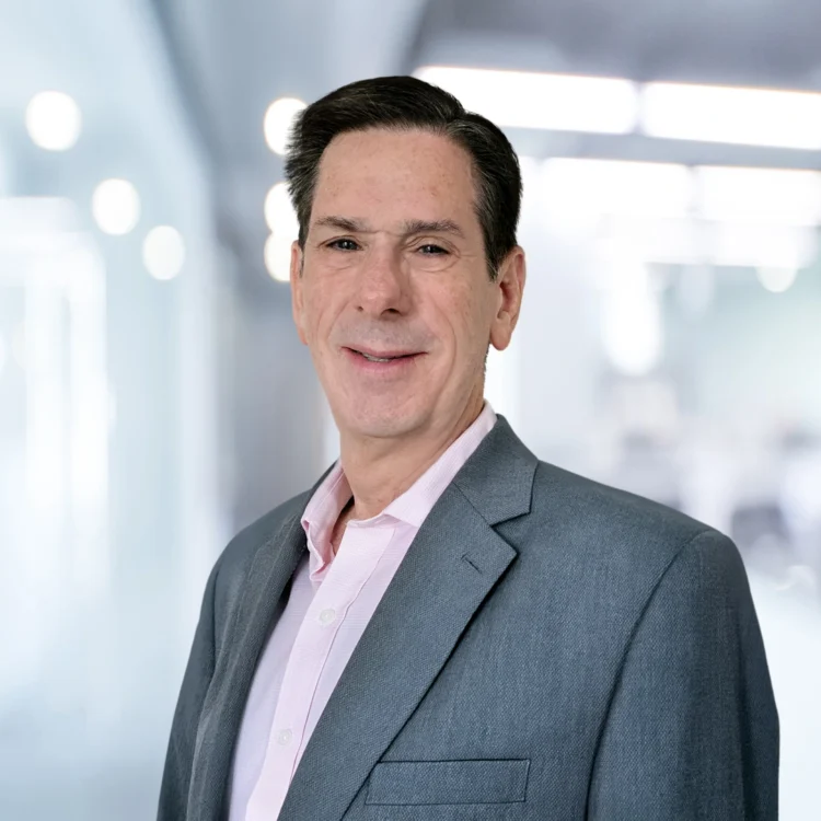 Man in a gray suit jacket and light pink shirt stands in a modern, brightly lit office environment, looking at the camera and smiling.