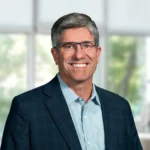A middle-aged man wearing glasses, a blue dress shirt, and a dark checked blazer smiles in front of a blurred window background.