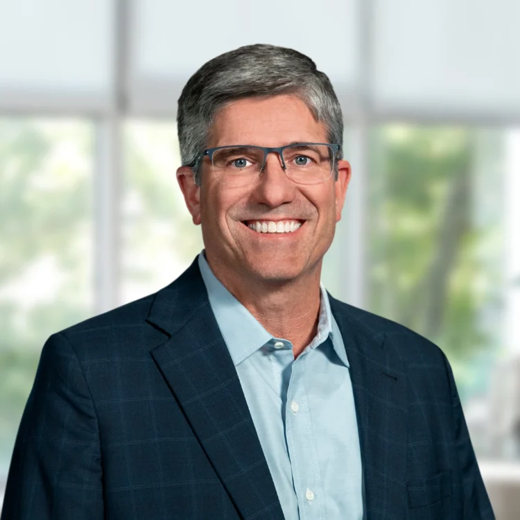 A middle-aged man wearing glasses, a blue dress shirt, and a dark checked blazer smiles in front of a blurred window background.