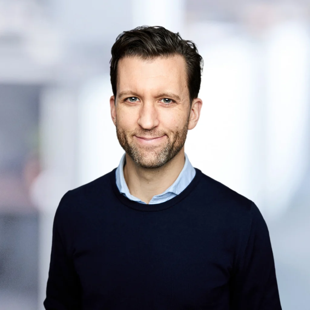 A man with short brown hair and a trimmed beard, wearing a navy sweater over a light blue shirt, stands in front of a blurred indoor background.