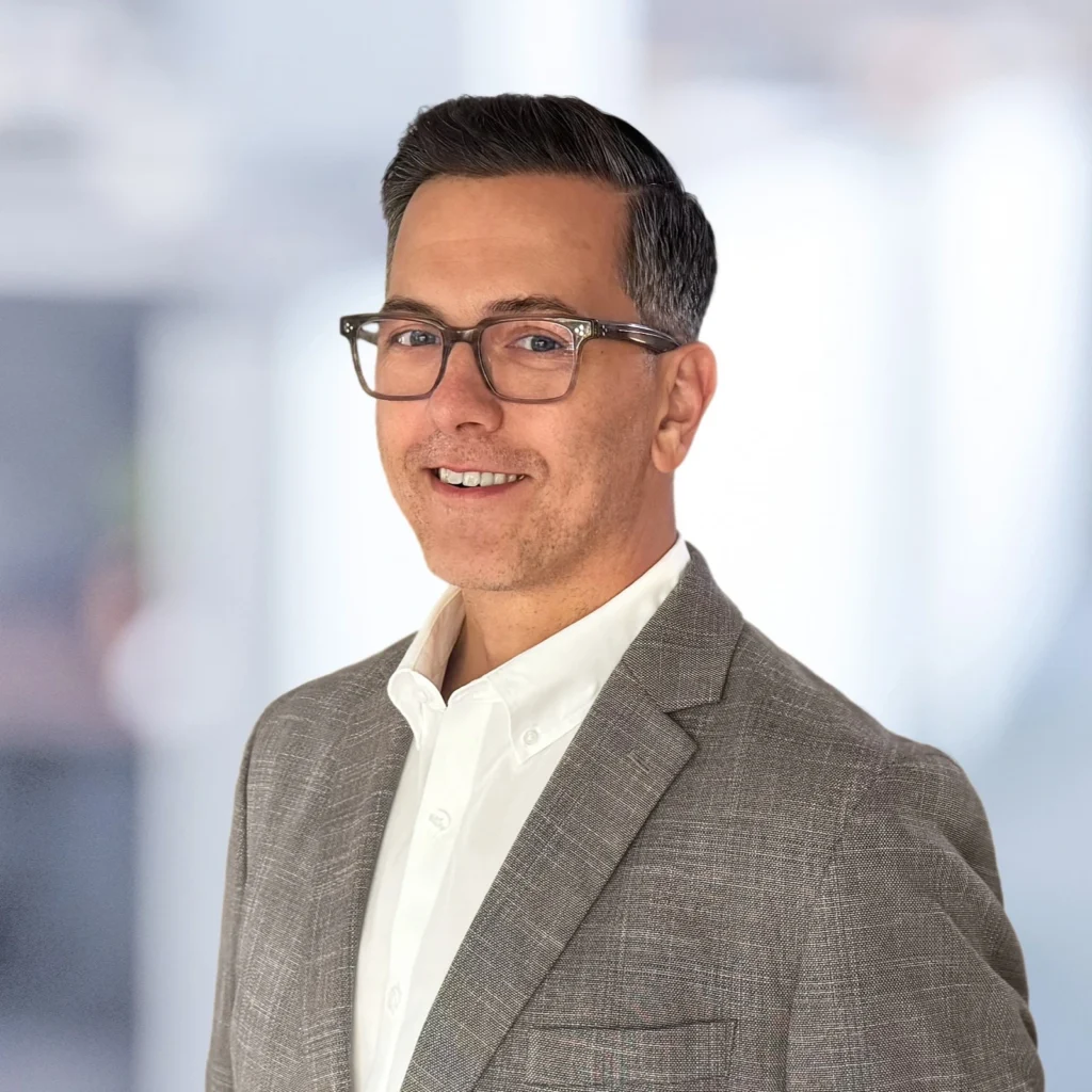 A man wearing glasses, a white shirt, and a gray blazer smiles while posing against a softly blurred indoor background.