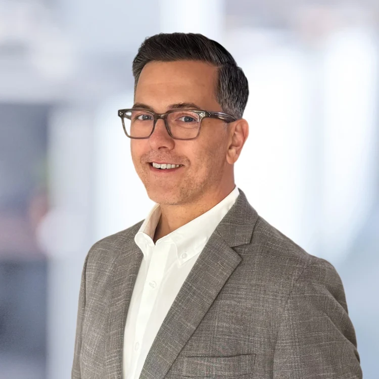 A man wearing glasses, a white shirt, and a gray blazer smiles while posing against a softly blurred indoor background.