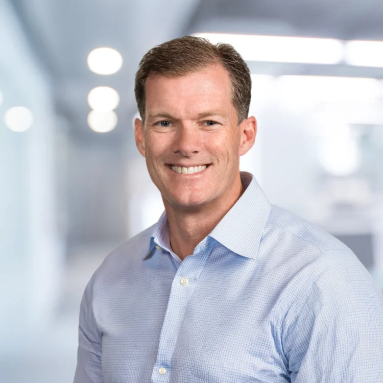 A man with short brown hair wearing a light blue button-up shirt is smiling, standing in a modern, brightly lit office environment.