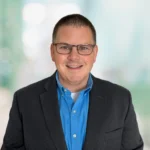 A man wearing glasses, a dark suit jacket, and a blue shirt smiles at the camera with a blurred light background.