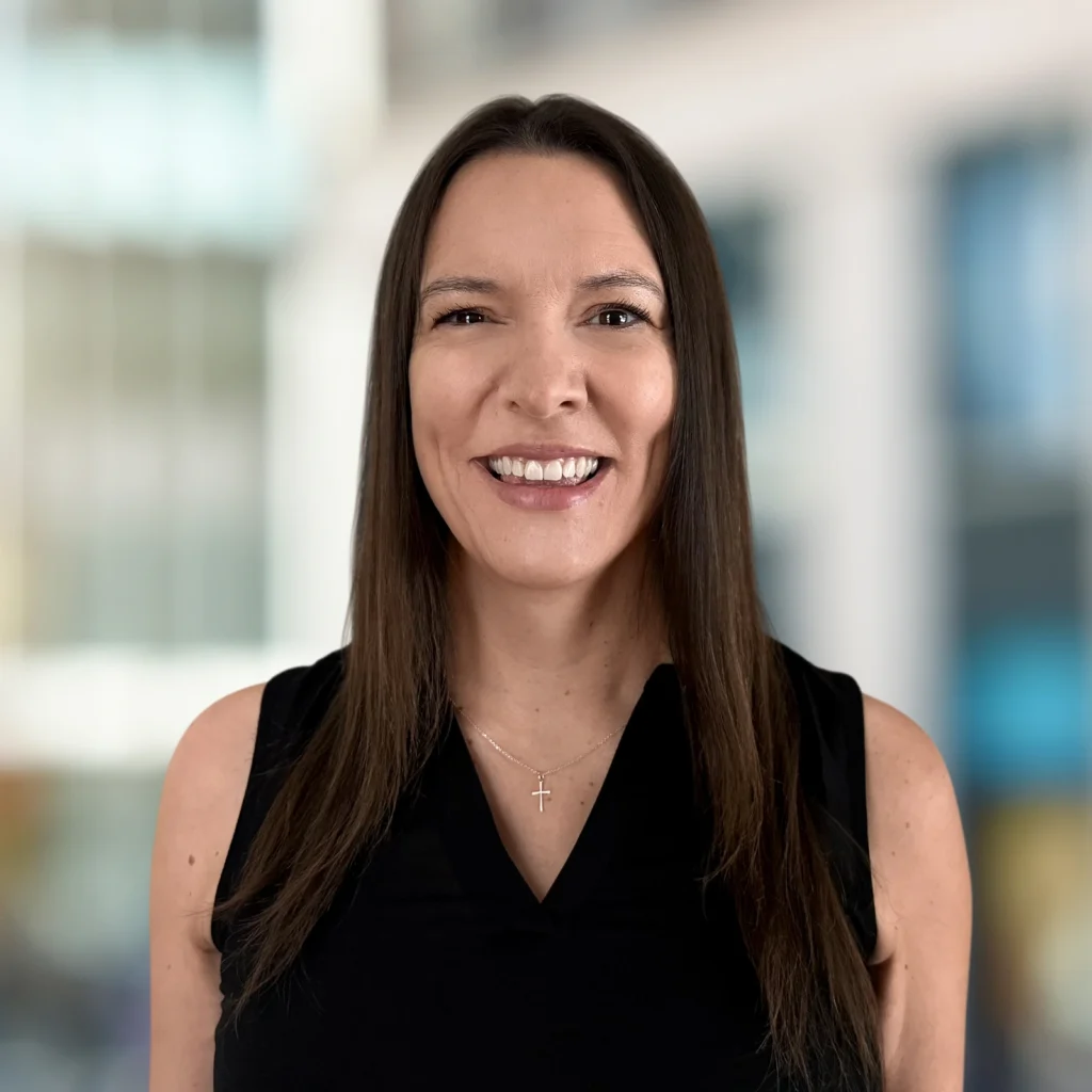 Woman with long brown hair, wearing a black sleeveless top and a cross necklace, smiling in front of a blurred urban background.