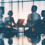 Four people sit around a table in an office, engaged in discussion, with laptops and papers in front of them and large windows in the background.