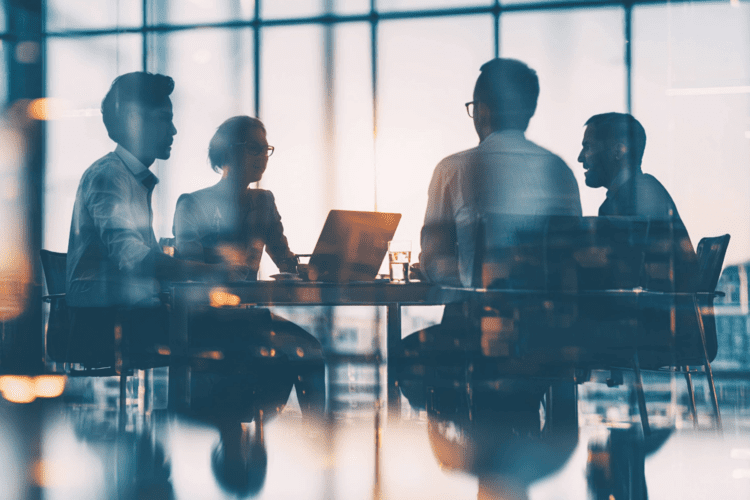 Four people sit around a table in an office, engaged in discussion, with laptops and papers in front of them and large windows in the background.