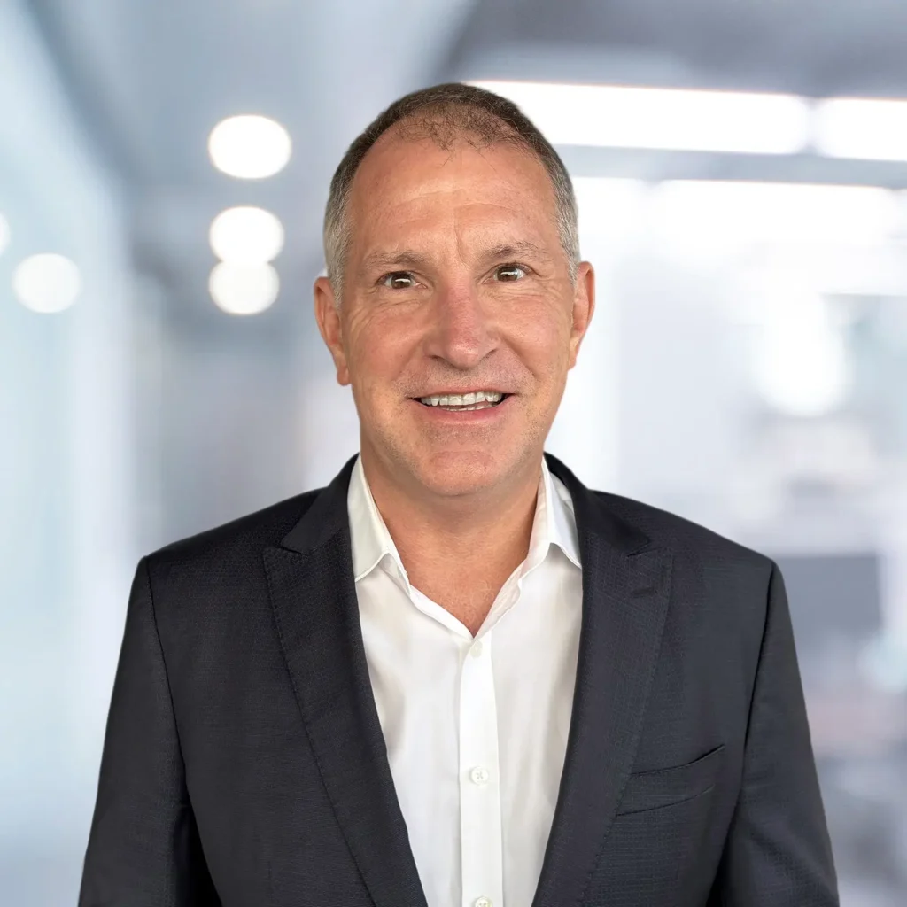 A middle-aged man in a dark suit and white shirt smiles while standing in an office setting with blurred lights in the background.