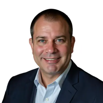 A man in a dark suit jacket and light blue collared shirt smiles at the camera against a plain white background.