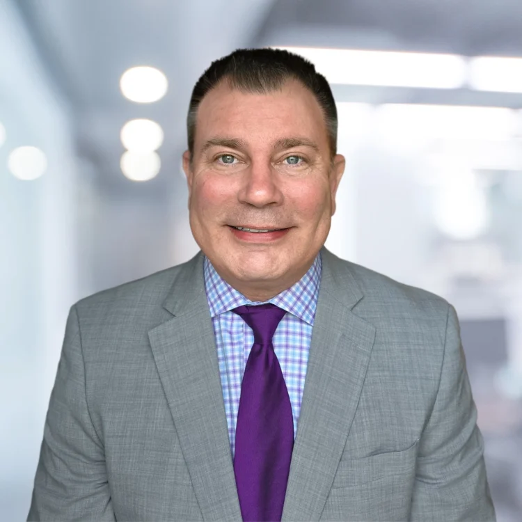 A man in a gray suit, purple tie, and checkered shirt smiles at the camera in an office setting with blurred lights in the background.