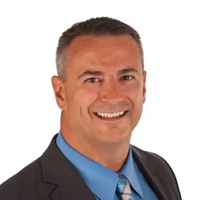 Smiling man with short dark hair wearing a dark suit jacket, blue shirt, and striped tie, posing in front of a plain white background.